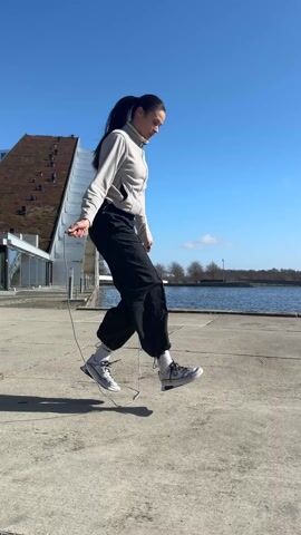 A young woman with long dark hair is jump-roping outdoors on a sunny day. She wears a light gray zip-up jacket, black pants, and light-colored sneakers. She faces right and jumps rope repeatedly. Behind her is a building with a white and silver architectural feature and a brown roof. In the background is a body of water under a clear blue sky. The area where she jumps is a concrete surface.
