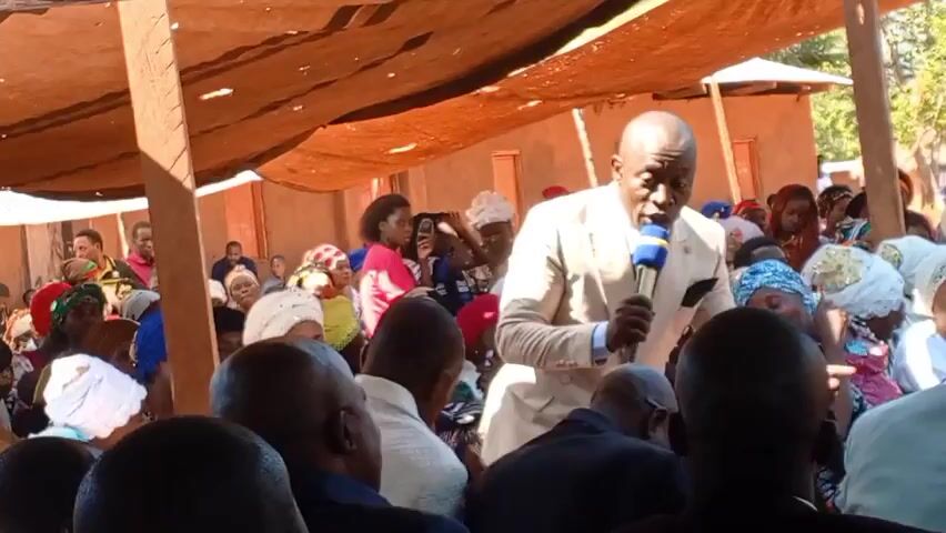 Here's a matter-of-fact description of the video:

A man in a light-colored suit is speaking into a microphone to a gathered crowd. The crowd appears to be mostly African, with many women wearing head coverings. They are seated under a makeshift awning, and some individuals are looking at the speaker. A few buildings and trees can be seen in the background. A person in the foreground on the right is holding a phone and watching the speaker. The speaker is animated, gesturing with his hands as he speaks. The scene seems to be taking place outdoors.