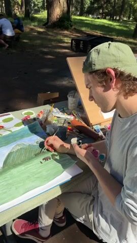 A young man wearing a green baseball cap and a light gray long-sleeved shirt is sitting at a table outdoors, painting a picture of a landscape. He holds a paintbrush in his right hand and a small palette with several colors in his left. The painting on the table depicts rolling green hills with some brown figures that appear to be bears. Various art supplies, including tubes of paint, brushes, and water cups, are spread across the table. In the background, a few other people are visible, some sitting down, and trees and foliage suggest a park or forest setting. The lighting indicates it is daytime.