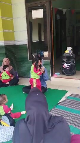 A young girl in a green shirt and pink pants stands holding a microphone and singing in front of an adult. She is on a green mat outdoors, in front of a building with a glass door. Several other children and adults are sitting on mats or the ground watching her. A large black speaker sits to the right of the girl. The adults are wearing hijabs.