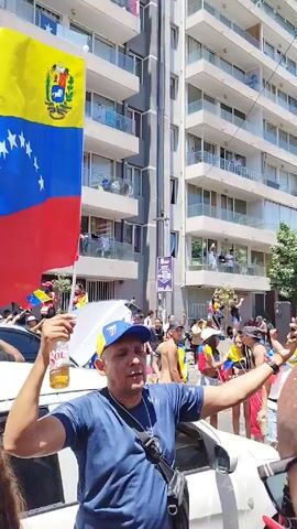 The video shows a daytime street scene with a crowd of people, many of whom are waving the flag of Venezuela. They appear to be gathered near or around several white cars. One man is holding a can of "Sol" brand beverage and waving a flag. The buildings in the background are modern apartment blocks with balconies. The atmosphere seems festive and patriotic.