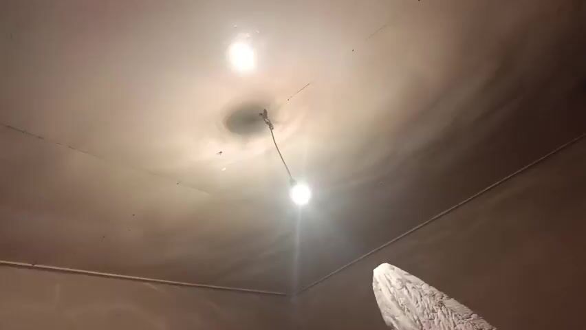 The video shows an interior room with plain, tan-colored walls and a textured ceiling. Two bare light bulbs are visible, one hanging from a wire in the center of the frame and another in the upper left corner. A white pipe runs along the ceiling line. In the lower right of the frame, a portion of what appears to be a white blanket or cloth is draped over an unseen object. The room contains a microwave oven on a stand, with a black appliance and electrical cords plugged into a wall socket above it. To the left, a partially open doorway reveals another room with dark shelving. A young boy wearing a yellow t-shirt with a dark logo is visible in the doorway, moving into the room and looking towards the right side of the frame. He appears to be laughing or smiling. The lighting is dim and the overall atmosphere suggests a casual, possibly domestic setting, possibly undergoing renovation or in a state of disarray.