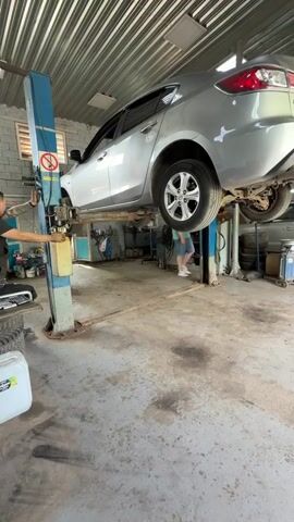 A silver sedan is raised on a hydraulic car lift in a garage. A man is standing to the left of the lift, operating a control panel with his hand. Another man is standing behind the car, near the rear passenger side. The garage appears to be cluttered with tools and equipment, including tires, canisters, and a toolbox. The floor is concrete and stained with oil or grease. The walls are made of cinder blocks, and the ceiling is corrugated metal.