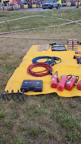 The video displays an outdoor scene, likely a preparation area for an event, with a focus on emergency equipment. A large yellow tarp is spread across a grassy area, serving as a staging ground for various tools and safety gear. Prominently featured are numerous red fire extinguishers, arranged in rows. Alongside them are power tools, including what appears to be a reciprocating saw and an impact driver, as well as extension cords, a toolbox, and other implements such as a shovel, wrenches, and what look like cutting tools. In the background, a crowd is visible, suggesting an audience or participants for an event. Further back, a vehicle and people in what might be uniforms are present, indicating ongoing activity. The overall impression is one of readiness and organization for a potentially hazardous situation or a competitive event.
