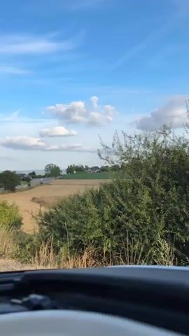 The video shows a rural landscape from the perspective of someone in a vehicle. The sky is blue with scattered white clouds. In the foreground, there is a patch of green bushes and dry grass. Beyond that, there is a field of golden-brown stubble, likely harvested grain. Further in the distance, there are more fields, trees, and some buildings, suggesting a rural settlement. A paved road with a white line marking its edge is visible, curving to the right. There are telephone poles with wires along the side of the road. The sun appears to be low in the sky, casting a warm light on the landscape. There are no people visible in the video. The vehicle appears to be moving along the road.