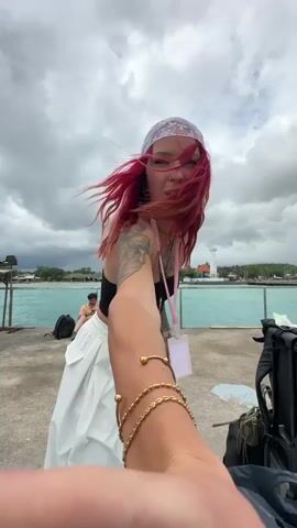 A person with red hair is recording a video of themselves on a concrete pier. They are wearing a black top, white baggy pants, and a white patterned headscarf. They have tattoos on their arms and are wearing gold bracelets. They are facing the camera and making dance moves. Behind them is a body of water, with a lighthouse visible in the distance. The sky is cloudy. Another person is sitting on the pier behind them, and luggage is visible nearby.