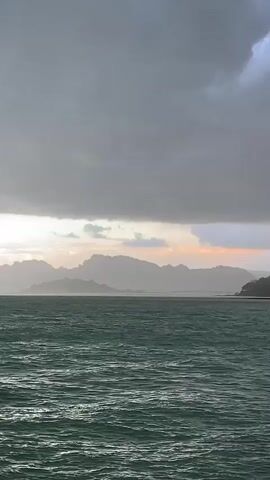 The media file captures a scenic view of the sea with a dramatic sky. The foreground is dominated by the dark, choppy water of the sea, reflecting hints of light. In the middle ground, a hazy silhouette of a distant mountain range stretches across the horizon, with what appears to be a bridge or structure visible in the far distance. The sky above is a striking composition of dark, heavy storm clouds that cover most of the frame, contrasted by a band of lighter, warmer-toned sky just above the mountains, suggesting either sunrise or sunset. The overall mood is one of nature's power and grandeur, with a sense of impending weather change. The location is identified as Don Sak, Thailand, which is known for its beautiful coastal scenery and often experiences dynamic weather patterns. There is no visible text in the media file.