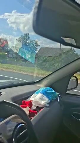 The video begins from the interior of a car, showing the dashboard and a portion of the windshield. Bags of items are visible on the dashboard. The camera then pans to the windshield, revealing a view of a road and the surrounding environment. A truck with some items on its flatbed is in front of the car. The car is driving on a paved road through a rural area with trees and houses visible on the sides. Another car is ahead on the road. The sky is blue with some clouds. The car continues to drive forward.