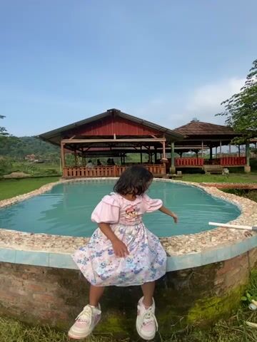 A young girl is sitting on the brick edge of a small, kidney-shaped pond or pool, dipping her hand in the water. She is wearing a pink and lavender dress with puffy sleeves, and white sneakers with pink details. She looks into the pool, then at the camera, smiling briefly. The water in the pool is turquoise. Behind her are two covered wooden structures with red roofs and railings. People are sitting inside the closest structure. Green grass surrounds the pool and the structures. Trees are visible in the background and a clear blue sky is overhead. The camera angle is low, looking slightly up at the girl.