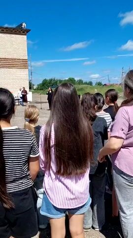 A group of young people, mostly girls with long hair, stand facing away from the camera, listening to two people speaking at a microphone in front of a beige brick building with a round tower. The sky is bright blue with scattered white clouds. A microphone is set up on a stand, and the speakers appear to be dressed formally, one in a black outfit and the other in lighter clothing. The audience members are casually dressed in t-shirts and shorts. The setting appears to be outdoors, possibly for an event or assembly.