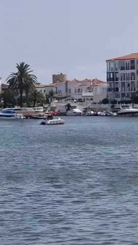 A person is seen on a small white boat with a red stripe on the side, moving through the water. The boat has an awning or canopy visible. In the background, there are several other boats docked and anchored, and a shoreline with white buildings and a palm tree. The sky is clear and blue.