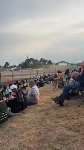 Here is a concise description of the video:

The video shows spectators sitting on a grassy hillside overlooking a racetrack. The racetrack is visible in the distance, with race cars speeding around the track. Some of the spectators are sitting in folding chairs, while others are sitting directly on the grass. Some are looking at their phones, while others are watching the race. The sky is overcast.