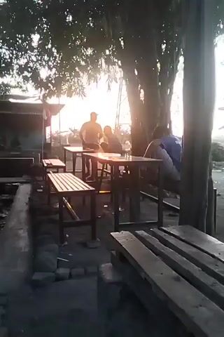 The video shows an outdoor seating area with wooden tables and benches. A large tree is prominently featured, with its branches casting shadows over the scene. The sun is setting, casting a warm, golden light and creating lens flare. Two people are visible, one sitting at a table and another person partially visible in the background. There is a small structure on the left side of the frame, possibly a shop or stall, and some debris on the ground. The overall atmosphere is relaxed and somewhat rustic.