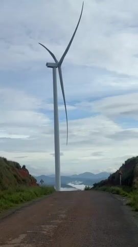 The video shows a wind turbine standing tall on a hilltop road. The road slopes downward, framed by grassy embankments on either side. In the distance, mountains rise above a layer of fog or clouds. The sky is partially cloudy, with patches of blue visible. A few road signs are visible at the edges of the road. The wind turbine has three blades and appears to be stationary.