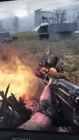 The video shows a first-person perspective from a video game. The player is holding a rifle with a crosshair and is near a fire. In the distance, there are concrete structures and a grassy field. There are at least two figures visible in the field. One is lying down, and the other is standing and appears to be shooting.