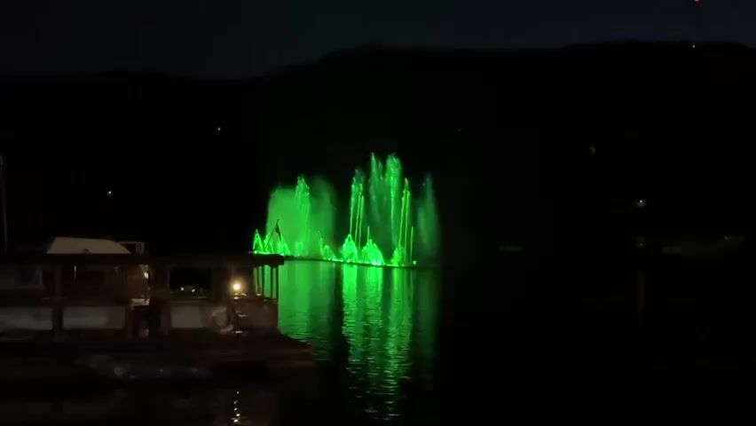 The video shows a nighttime scene on a body of water. In the center of the frame, a fountain is illuminated with bright green lights, creating a dramatic visual effect on the dark water. The fountain jets appear to be in motion, with streams of water shooting upwards. In the foreground on the left, there is a dock or a boat structure with some lights visible, casting reflections on the water. In the background, a dark silhouette of hills or mountains is visible against the night sky. The overall atmosphere is one of nighttime entertainment or a light show.