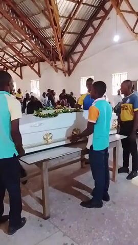 Here is a forensic description of the video:

The video shows the inside of a church-like building. A white coffin, adorned with floral arrangements, rests on a wooden table. The coffin has gold-colored handles. Around the coffin, several men stand, some wearing turquoise and orange shirts, others in blue and yellow. These men appear to be pallbearers, as they are positioned to lift and carry the coffin. Seated behind the coffin, out of focus, are a number of people who appear to be mourners. The interior of the building has a high, unfinished-looking wooden roof structure with open beams. There are windows on the right side of the frame, letting in natural light. The floor appears to be a light-colored tile or linoleum. The camera seems to be hand-held and provides a static shot, capturing the somber scene of what appears to be a funeral service.