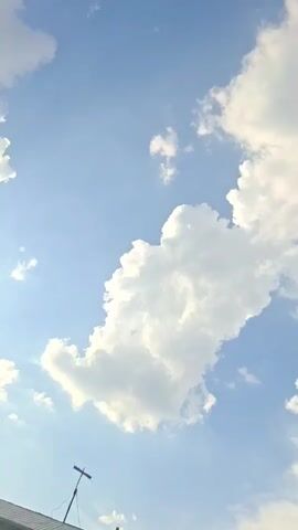 This is a vertical, close-up shot of a clear blue sky with large, fluffy white clouds. The clouds are illuminated by sunlight, creating a bright and airy atmosphere. In the lower left corner of the frame, the top of a building with a rooftop antenna is visible. The camera is pointed upwards, capturing the vast expanse of the sky.