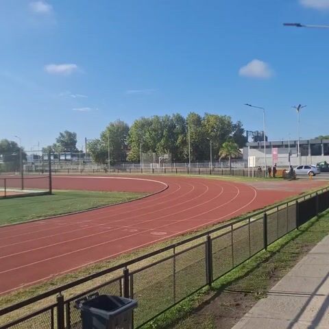 The video shows a sports field on a sunny day with a blue sky dotted with fluffy white clouds. The video begins showing the curve of a red running track surrounding a green sports field. A black trash bin stands near a dark metal fence in the foreground. As the camera pans across the field, it shows a soccer goal, benches, and light poles surrounding the field.