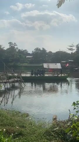 A ferryboat with a green hull and a white roof is crossing a body of water. There are several people standing on the deck of the ferryboat. On the left side of the image, there is a wooden dock extending into the water, with a ramp leading up to the ferryboat. The ferryboat is decorated with a red and white flag. The water is calm and reflects the sky and surrounding trees. The background shows a lush green landscape with trees and some buildings. The sky is partly cloudy with sunlight breaking through. The overall scene depicts a peaceful and scenic river crossing.