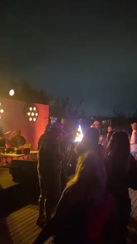 The video shows a group of people gathered outdoors at night. There is a stage with lighting and what appears to be musical equipment. Several people are standing and talking, while others are closer to the stage, possibly watching a performance. The lighting is dim and colorful, creating a party atmosphere. The ground is wooden decking. The sky is dark, with some clouds visible.
