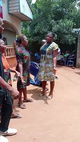 The video shows several people milling around in an outdoor setting, likely a yard or gathering space outside a building. The ground appears to be a mix of dirt and sand, and the overall ambiance suggests a community event or gathering. The people are dressed in varied clothing, some in what appears to be traditional or patterned attire, while others are in more casual wear. People are walking and moving around, and their interactions are unclear without sound. A building is visible in the background, and other details, like chairs and possible event-related items, are scattered throughout the scene.