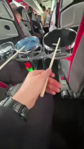 The video shows a person playing a small drum set on a bus. The drum set consists of three drums of varying sizes. The person is holding drumsticks and striking the drums. The bus is full of passengers, and the seats are red and gray. The person is wearing a black shirt and a watch. The drum set is positioned between two seats, partially blocking the aisle.