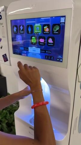 A person's arms are visible from the chest down, with a woman's hands and arms wearing a pink bracelet and a red bracelet, interacting with a white Bevi beverage dispenser. The dispenser has a large touch screen displaying various drink options with fruit icons. The woman's left hand is holding a clear plastic bottle, positioning it under the dispenser spout. Her right index finger is pressing the "POUR" button on the touch screen. The dispenser's interior light illuminates the bottle with a blue glow. The background is slightly out of focus, showing some greenery to the left and part of another white structure to the right. The overall environment appears to be indoors.