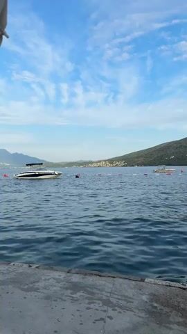 The video shows a view of a body of water from a concrete surface. The water is blue and has small waves. In the distance, there are green hills with some buildings on them, and a few boats. The sky is blue with some white clouds. A white and blue speedboat is visible in the middle of the frame, and another boat is further in the distance on the right. There are buoys on the water, marking a boundary. The concrete area in the foreground appears wet, possibly from the waves or recent rain. The overall scene is calm and scenic, suggesting a coastal or lakeside location.
