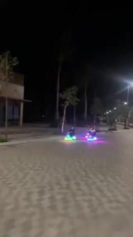 At night, two small, brightly illuminated amusement ride vehicles are driven on a paved walkway. The rides appear to be similar to bumper cars, but smaller, and have neon lights in multiple colors. They are each driven by at least one person. The vehicles are driven on a walkway with patterned pavement and palm trees along the edge. Buildings are visible in the background. The person driving the ride on the left waves to the camera.