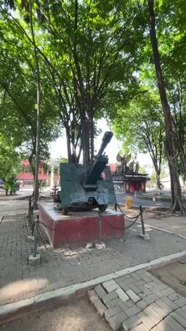 A stationary video shows a large, green artillery cannon mounted on a red pedestal. The cannon is positioned in an outdoor area with cobblestone paving. Tall trees with lush green foliage surround the cannon, creating a shaded environment. In the background, there are buildings and a monument with what appears to be eagle statues. The overall scene suggests a public space, possibly a park or a memorial site.