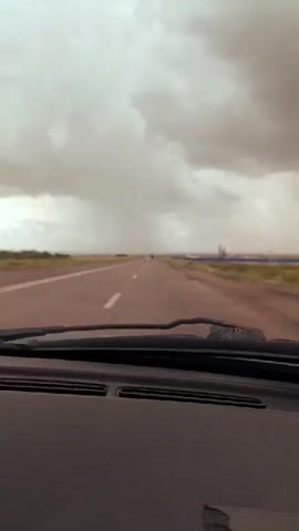The video captures a view from inside a car, looking forward through the windshield at a straight, empty highway stretching into the distance. The sky is heavily overcast with dark, ominous clouds, suggesting impending rain or a storm. In the far distance, there appears to be a light shower falling on the road. The landscape on either side of the highway is flat and appears to be rural, with vegetation visible. The dashboard of the car is visible in the foreground.