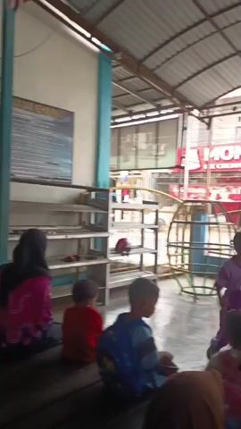 The video shows a group of young children, most wearing purple uniforms, gathered in an open-air covered area. The children appear to be engaged in some sort of activity, possibly a dance or game, as several are moving their bodies and interacting with each other. Some children are seated, watching the others. There is a playground structure in the background, along with shelves and a screen displaying an image. In the far background, there are buildings and what appears to be a street. The ground is wet, suggesting it may have recently rained. The overall atmosphere appears to be one of a school or daycare setting.