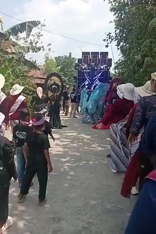 A group of people are gathered on a paved road. In the background, a large sound system with speakers is set up. In the center of the road, a person dressed in a sparkly blue costume with large wings is standing. Two circular decorative frames, one black and one red, are positioned on either side of the person in the blue costume. Several individuals, including children, are observing the scene. Some of the observers are wearing hats and traditional clothing. The environment appears to be outdoors with trees and houses visible. The scene suggests a festive or celebratory event.