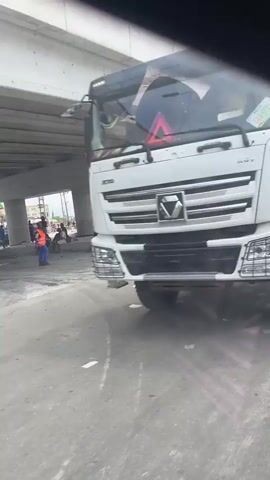 The video captures a dynamic street scene in Effurun, Nigeria, from the perspective of a moving vehicle. It opens with a close-up of a large white truck's tractor unit, displaying the partially visible text "ECL/TRT1029" on its side.

The camera then pans to reveal a wide shot of a multi-lane road passing beneath a substantial concrete overpass or bridge, supported by numerous pillars. Several individuals are present: pedestrians walk along the roadside, and groups of people are gathered or standing under the bridge structure, some seemingly engaged in conversation. A white van with passengers, including children looking out of the windows, passes by. Other vehicles spotted on the road include a motorcycle, a blue tricycle (keke napep), and a silver car in the distance.

Notable details include a significant amount of litter and debris scattered on the ground along the curbs and under the bridge. Red and white barrier tape is also visible under the overpass, possibly marking an area or indicating construction/hazard. The scene takes place during the day under what appears to be a clear, bright sky.