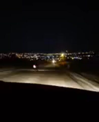 The video shows a nighttime scene, likely from the perspective of someone in a vehicle. The foreground is dark, with the headlights of the vehicle illuminating a dirt road. In the distance, a city skyline is visible, dotted with lights. A lone motorcyclist is also visible on the road, their headlights shining brightly. The overall atmosphere is one of quiet nighttime travel, with the distant city lights providing a sense of destination or activity. The location is identified as Bahşılı Belediyesi, Turkey. The time of day is clearly night, and the weather appears to be clear, allowing for the visibility of the distant city. There is no visible text in the video.