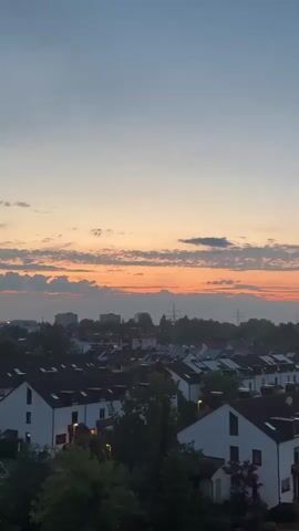 The video shows an aerial view of a residential neighborhood at dusk. The sky is a gradient of blue, with a band of orange and pink clouds near the horizon, indicating sunset. Below the sky, rows of white houses with dark roofs are visible, interspersed with trees. The houses appear to be townhouses or terraced houses. Some streetlights are illuminated, casting a warm glow on the houses and pathways. The overall impression is a peaceful evening scene in a suburban area.