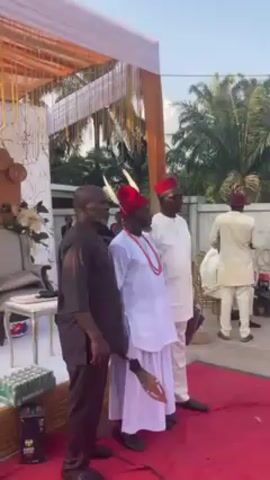 Here is a detailed description of the video:

The video appears to capture a traditional or cultural event taking place outdoors. Several men are dressed in traditional clothing. Some wear white outfits, traditional red caps, and red beaded necklaces. One man holds a ceremonial spear. A red carpet runs in front of a white and gold decorated area. There is a stack of Heineken beer visible as well.

Later in the video, a man dressed in a light-colored traditional outfit and red cap is seen standing in front of a tent that has a sign that says "NNokwa United Brothers" on it. A person in a black jacket approaches him. Other people wearing red caps are seated in the background.  The surrounding environment includes palm trees and other greenery. The video seems to be taken during the day.
