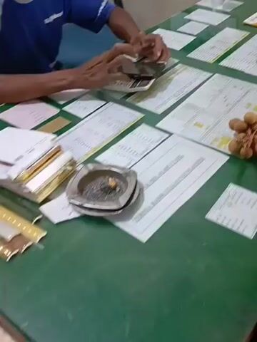 A medium shot shows a green table covered with various documents, papers, and a few personal items. In the foreground, a silver ashtray filled with cigarette butts sits on a document. To the left, a stack of papers is neatly piled. In the background, a person wearing a blue shirt is seated and appears to be using a calculator. To the right, another person wearing a pink hijab and glasses is seated, holding a small object in their hands. The environment suggests an office or a workspace.