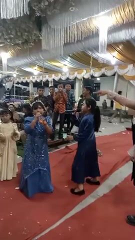 The video shows a group of children and adults gathered indoors, possibly at a celebration or event. The setting appears to be decorated with draped white and gold fabric overhead and long white strands hanging down. In the foreground, two young girls are the focal point. The girl on the left is wearing a sparkly blue dress and holding a microphone, singing or speaking into it. She is standing on a red carpet. To her right, another girl in a dark blue dress is facing her, also on the red carpet. Further back, several boys in casual clothing are clustered together, some looking towards the girls. An adult is visible behind the boys, partially obscured. To the far left, a young girl in a pale gold dress stands looking towards the right. Several other people are seated or standing in the background, blurred due to the camera's focus. Confetti or small pieces of paper are scattered on the red carpet. The lighting is artificial and appears bright.