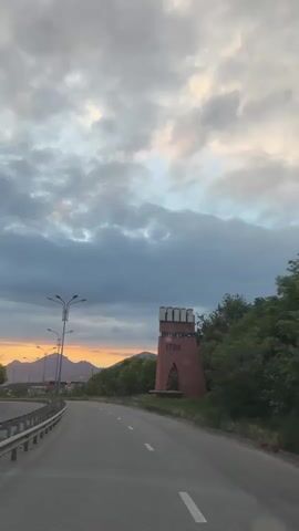 The media file captures a scenic view of a road leading towards mountains under a dramatic, cloudy sky at sunset. The setting appears to be Goryachevodsky, Russia, given the contextual information.

**Subjects:**
*   **Objects:** A distinct, reddish-brown brick tower with a distinctive shape featuring a triangular base and a crenellated top is prominently featured on the right side of the road. The number "1780" is clearly visible in large black numerals on the tower, suggesting a historical significance or a founding year. Streetlights with multiple lamps line the left side of the road, and a metal guardrail separates the road from an embankment. Lush green trees and vegetation border the road and surround the tower. In the distance, mountains are silhouetted against the sky.
*   **Scene/Setting:** The scene is a paved road curving gently to the right, suggesting it's a route into or out of an area. The presence of streetlights and a well-maintained road indicates it's likely a well-traveled path, possibly a highway or main road. The natural landscape with mountains and trees evokes a sense of rural or semi-urban outskirts.

**Activity or Event:**
The media appears to be a recording of a drive or a static view from a vehicle moving along this road. There's no discernible human activity, and the focus is on the landscape and the architectural element of the tower.

**Notable Details:**
*   **Time of Day:** The warm hues of orange and yellow peaking through the clouds indicate it is sunset, creating a dramatic and atmospheric sky.
*   **Weather:** The sky is heavily clouded, with dark gray clouds dominating, but breaks of lighter, illuminated clouds suggest the sun is low on the horizon.
*   **Location Cues:** While the general landscape points to a mountainous region, the specific mention of Goryachevodsky, Russia, provides definitive location context. The tower itself, with its inscribed year, is a significant landmark.
*   **Emotions:** The combination of the dramatic sky, the approaching dusk, and the striking historical tower can evoke feelings of contemplation, awe, or a sense of journey.

**Visible Text:**
The most prominent visible text is the large number "1780" on the brick tower. There might be additional text on the tower, but it is not clearly legible in this media.