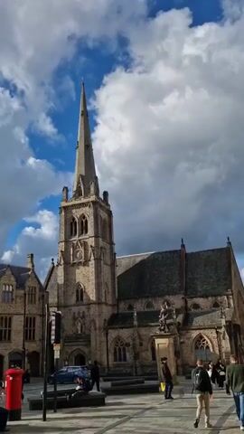 The video shows an outdoor urban scene. The sky is partly cloudy, with a mix of blue sky and gray clouds, and the sun occasionally peeks through the clouds. The camera pans around a square or plaza featuring historical buildings, including a church with a tall spire, and a building with an ornate facade labeled "Market Hall." There is also a statue of a person on horseback, and a red British-style post box. Several people are visible, including some sitting on benches, and others walking through the square. The overall impression is of a bustling town square on a partly cloudy day.
