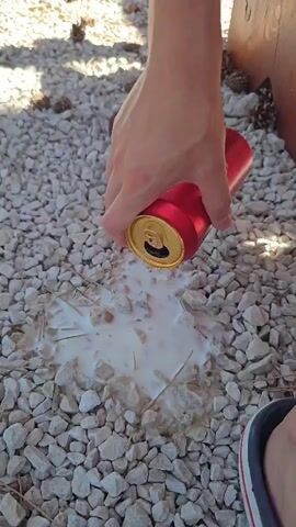A person's hand is shown tilting a red aluminum can, pouring a light-colored foamy liquid onto a bed of light-colored gravel. The liquid spreads out forming a puddle with some pieces of gravel mixed in. A portion of a person's foot wearing a sandal is visible in the lower right corner of the frame.