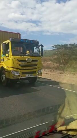 The video shows a view from a moving vehicle on a paved road. In the first scene, the camera focuses on a large yellow truck with a red stripe along the bottom. The video then shifts to show a white and blue bus with the words "THE GUARDIAN ANGEL" written on its side, passing in the opposite direction. The final scene shows a white SUV driving in the opposite direction, with the license plate "KDR 672E". The surrounding environment is a rural landscape with dry vegetation, hills, and a blue sky with scattered clouds. Utility poles are visible along the roadside.