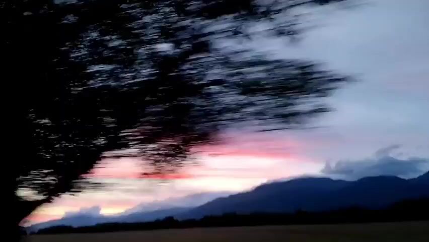 This video captures a fast-paced drive through a rural landscape during what appears to be either sunrise or sunset. The camera is mounted inside a vehicle, and the motion blur is significant, suggesting a high speed.

The foreground is dominated by the dark silhouette of trees and foliage, which whip past the frame. Through the gaps in the trees, a dramatic sky is visible. The sky displays a gradient of colors, ranging from deep blues and purples at the top to vibrant pinks and oranges closer to the horizon. Wispy clouds are scattered across the sky, catching the light and adding to the atmospheric effect.

In the background, the rounded, dark shapes of mountains or large hills can be seen, partially obscuring the horizon. The overall impression is one of dynamic movement and a fleeting glimpse of a beautiful natural scene. No people or specific objects are discernible due to the extreme motion blur. The environment appears to be natural and undeveloped, with hints of fields or plains in the lower portion of the frame.