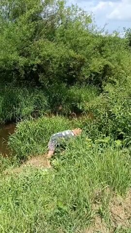 A person wearing a striped shirt and patterned shorts is crouched down in a grassy area next to a narrow stream. The person appears to be reaching into the tall grass. The stream is surrounded by lush green vegetation, including trees and tall grasses. The sky is partly cloudy.