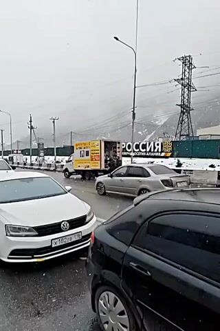 The video shows a street scene in what appears to be a mountainous region on a grey, snowy day. The road is wet and there are cars parked and driving along the street. There's a large sign reading "РОССИЯ - СЕВЕРНАЯ ОСЕТИЯ-АЛАНИЯ" and a truck labeled "ФРУКТЫ/С" parked near it. Several people are near the back of the truck. A white bus with "ТДТ СМО ТУР" written on its side drives past. The surrounding area includes power lines, buildings, and snow-covered mountains in the background.