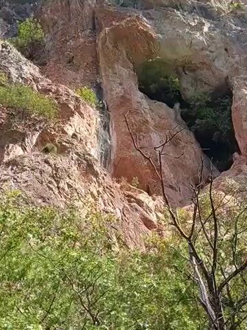 The video pans across a rocky mountainside covered in sparse vegetation. The camera moves from a close-up of the textured rock face, showing layers and crevices, to a wider view that reveals more of the mountain's slope and surrounding greenery. In the distance, a vast landscape of rolling, forested mountains stretches under a clear blue sky. Sunlight illuminates the scene, casting shadows and highlighting the rugged terrain.