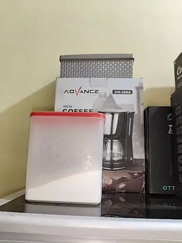 The video shows a shelf with several kitchen appliances and storage containers. On the left, there is a clear plastic container with a red lid, filled with a white substance, possibly sugar or flour. Behind it, there are boxes of coffee makers and a small appliance. To the right, there is a box for a blender. The items are arranged on a dark surface, possibly a countertop or shelf, against a light-colored wall. The lighting is soft and diffused, suggesting an indoor environment. There are no people or animals visible in the video.