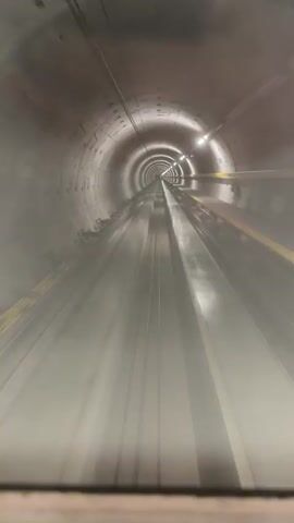 The video shows a first-person view of something moving rapidly through a circular tunnel. The tunnel is constructed from concrete segments, and the lighting is dim, with periodic lights along the ceiling. There are metal rails or supports running along the floor of the tunnel. The movement is smooth and steady, giving the impression of a mechanical system in operation. Audio includes speech.
